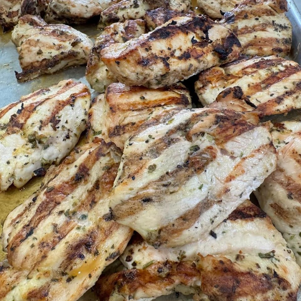 Close-up of numerous grilled chicken breasts with char marks and herbs on a metal tray.